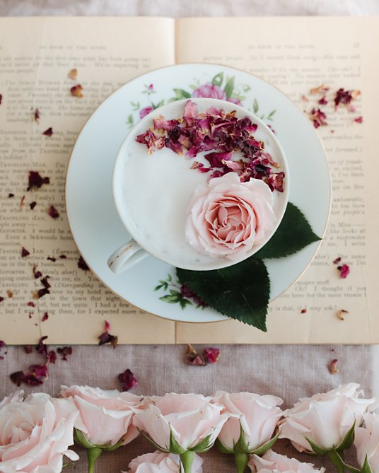 An artistic flat lay photo of a flower and flower petals in a teacup filled with white liquid and surrounded by decorations
