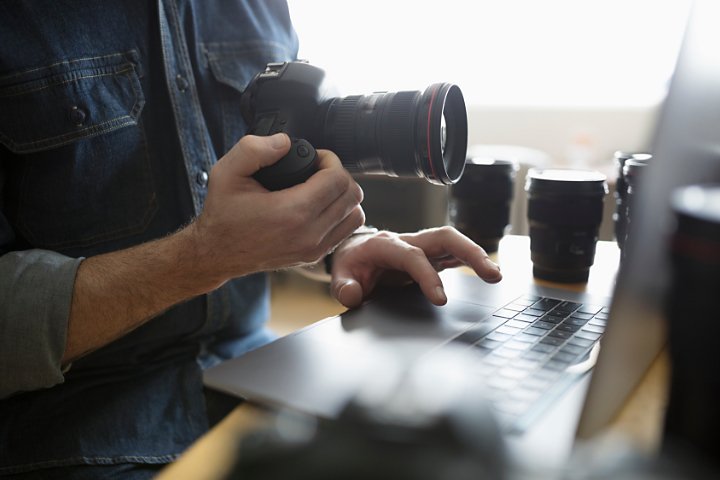 A photo of a person with a camera in one hand, using a laptop with their other hand.