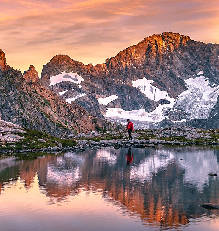 A rocky mountain range reflected in a body of water as a hiker travels next to it