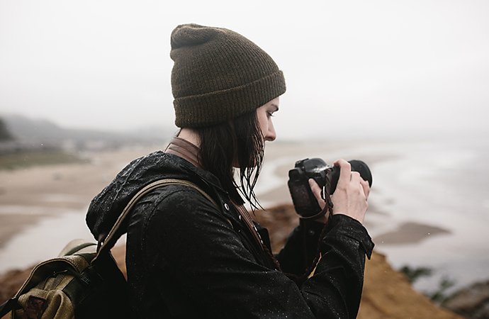 A mobile photo of a photographer looking at their DSLR camera while standing on a coastline