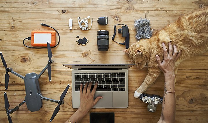 Aerial view of a desk with a drone, laptop, camera equipment, and an orange cat lying on it