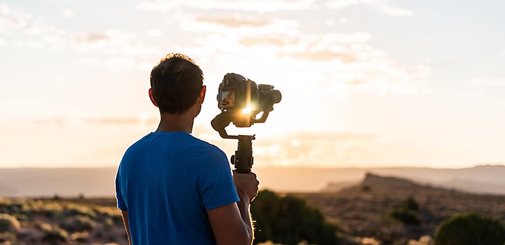 A photographer standing in front of a sunset holding a camera on a tripod
